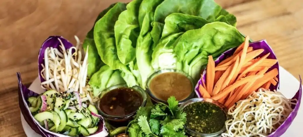 A white plate arranged with red cabbage leaves filled with lettuce, carrots, bean sprouts, cucumber, and three different dressings to dip them in.