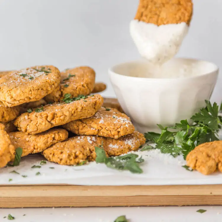 Pile of chickpea nuggets next to a bowl of vegan mayo ranch dip with a dipped nugget hovering over the bowl.