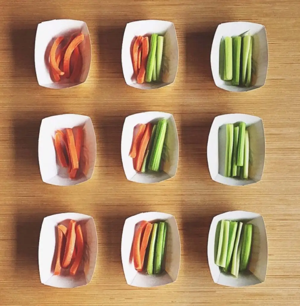 Nine white paper containers holding carrots and celery arranged in sets of three across on a wooden table.