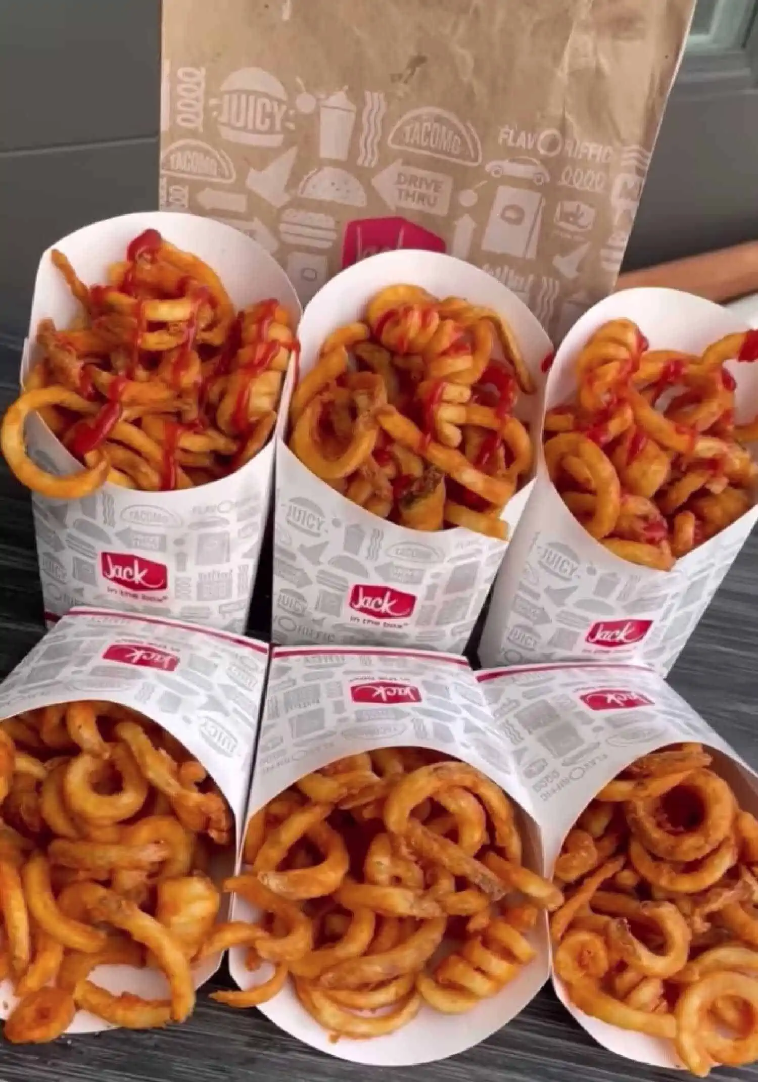 Six white boxes of Jack in the Box curly fries (some with ketchup) next to a brown take out bag from the restaurant.