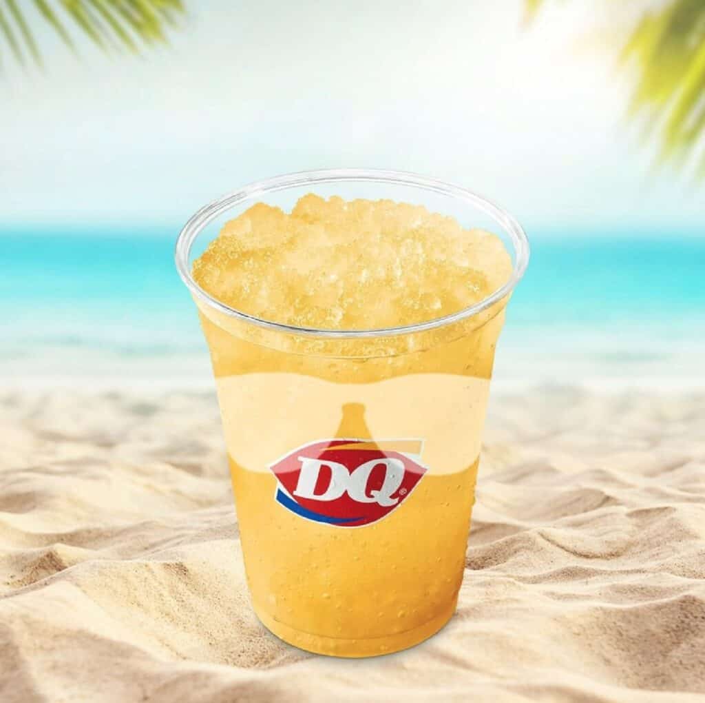 A sunny beach scene with a clear plastic cup filled with mango slush sitting on the sand. 