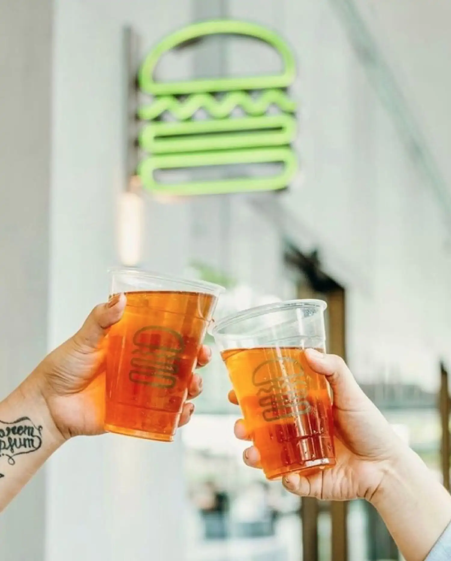 Two hands each holding a clear plastic cup filled with Shake Shack iced tea in front of a neon green Shake Shake sign.