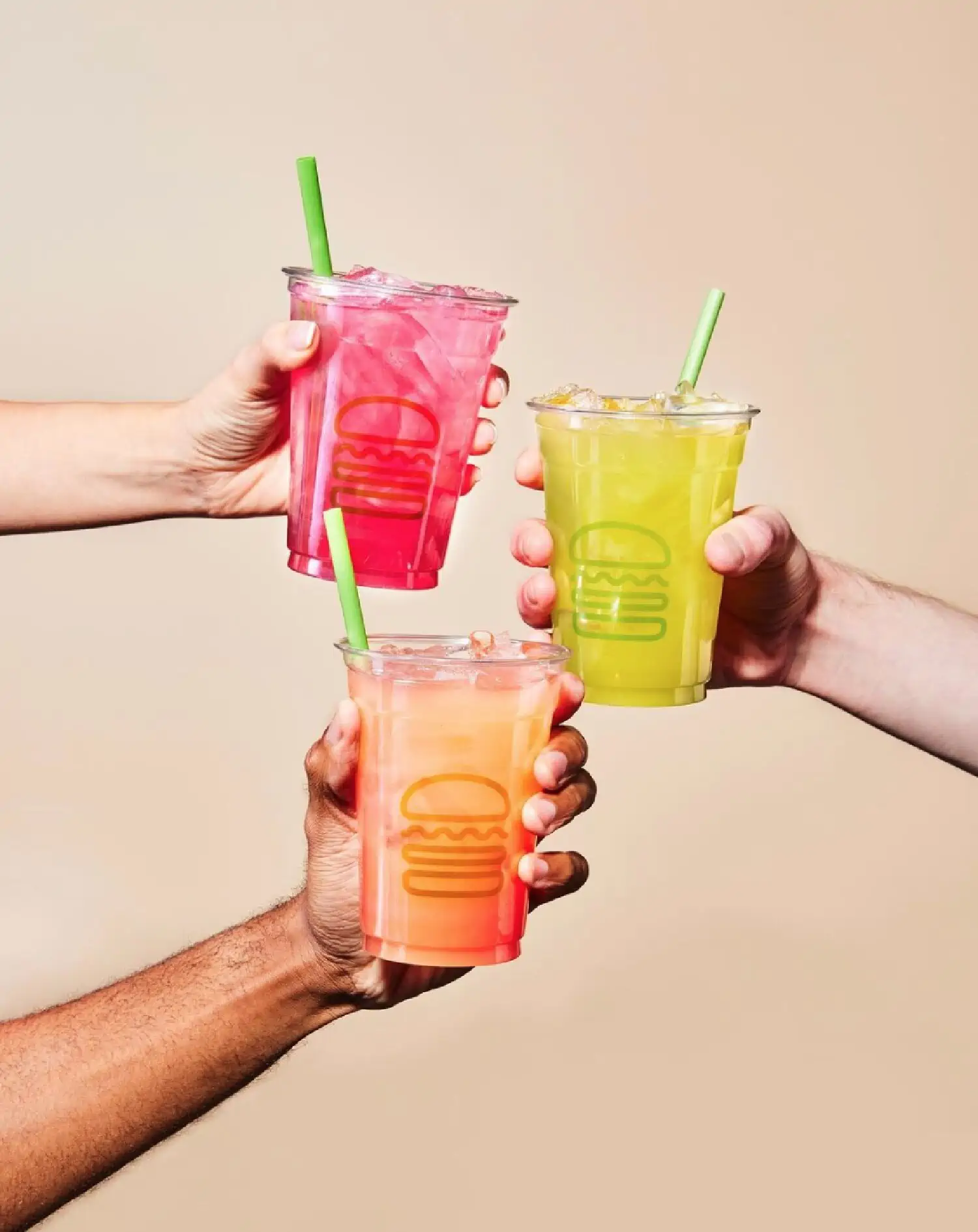 Three hands each holding a Shake Shack vegan-friendly fruit lemonade (Cherry Hibiscus, Mango Passionade, and Strawberry Lemonade) with a green straw against a beige wall. 