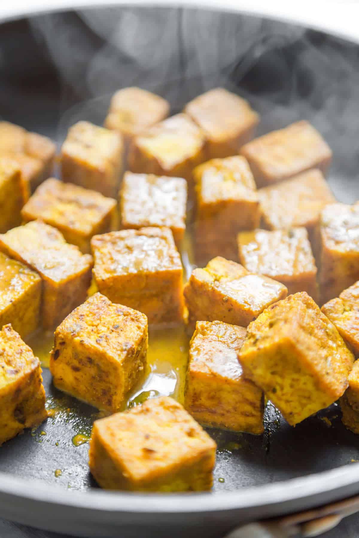 Tofu cooking in a large skillet with steam coming off.