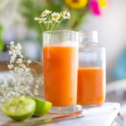 Tropical carrot juice in a glass next to a jar of carrot juice with lime wedges and flowers beside it.