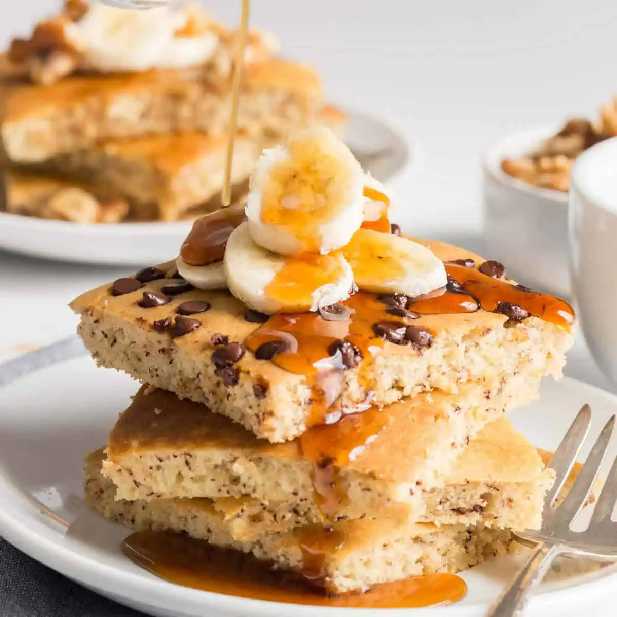 Vegan sheet pan pancakes stacked on a plate, topped with banana slices and maple syrup.