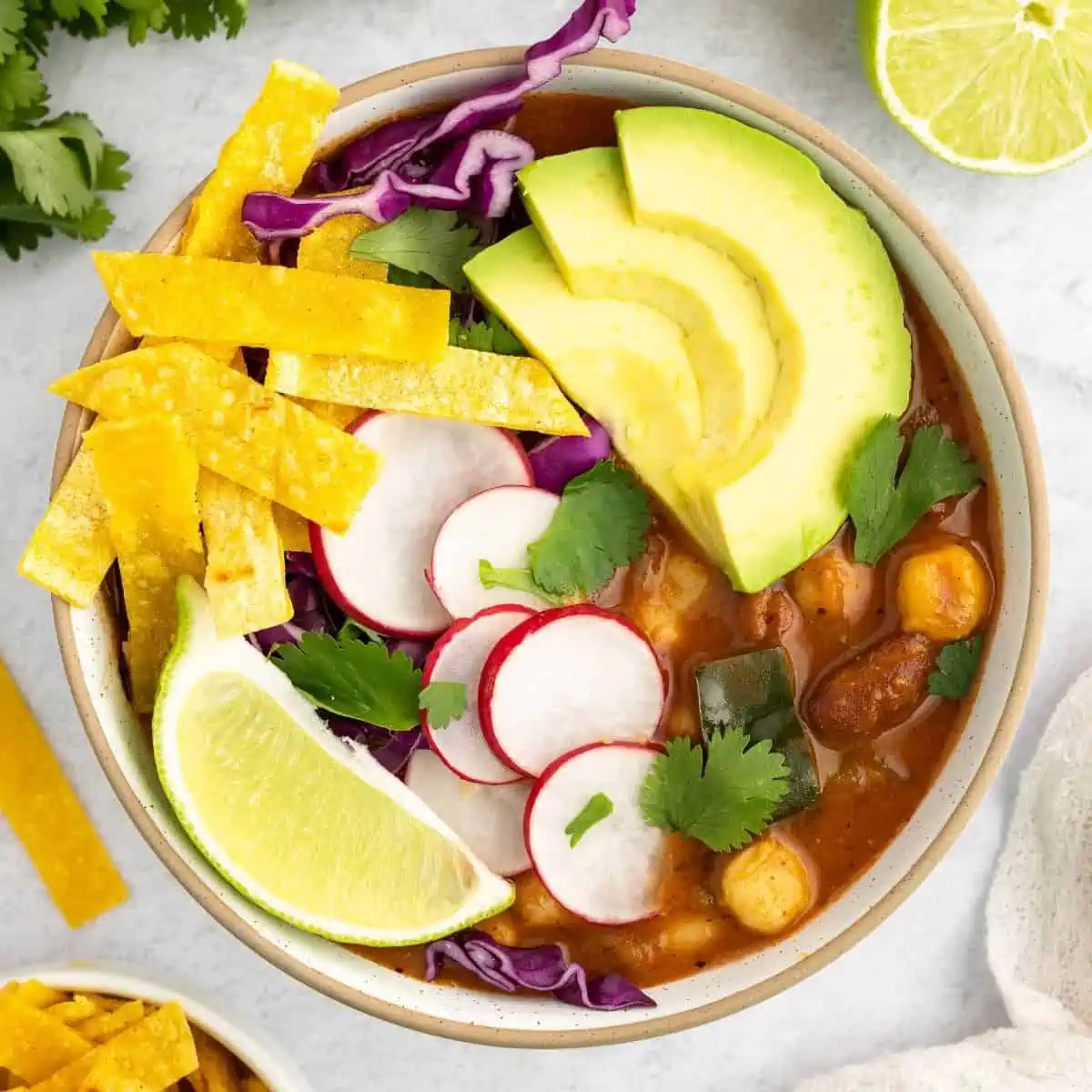 A bowl of vegan pozole with avocado, a lime wedge, radishes, and tortilla strips.