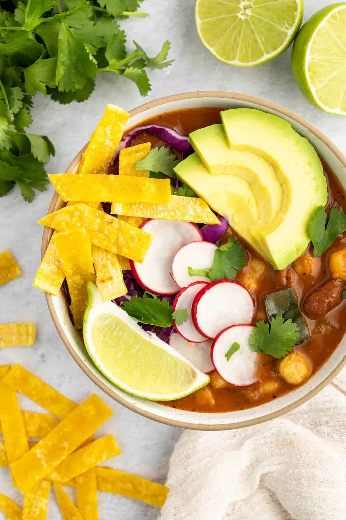 A bowl of vegan pozole with avocado, radishes, tortilla strips, and a lime wedge.