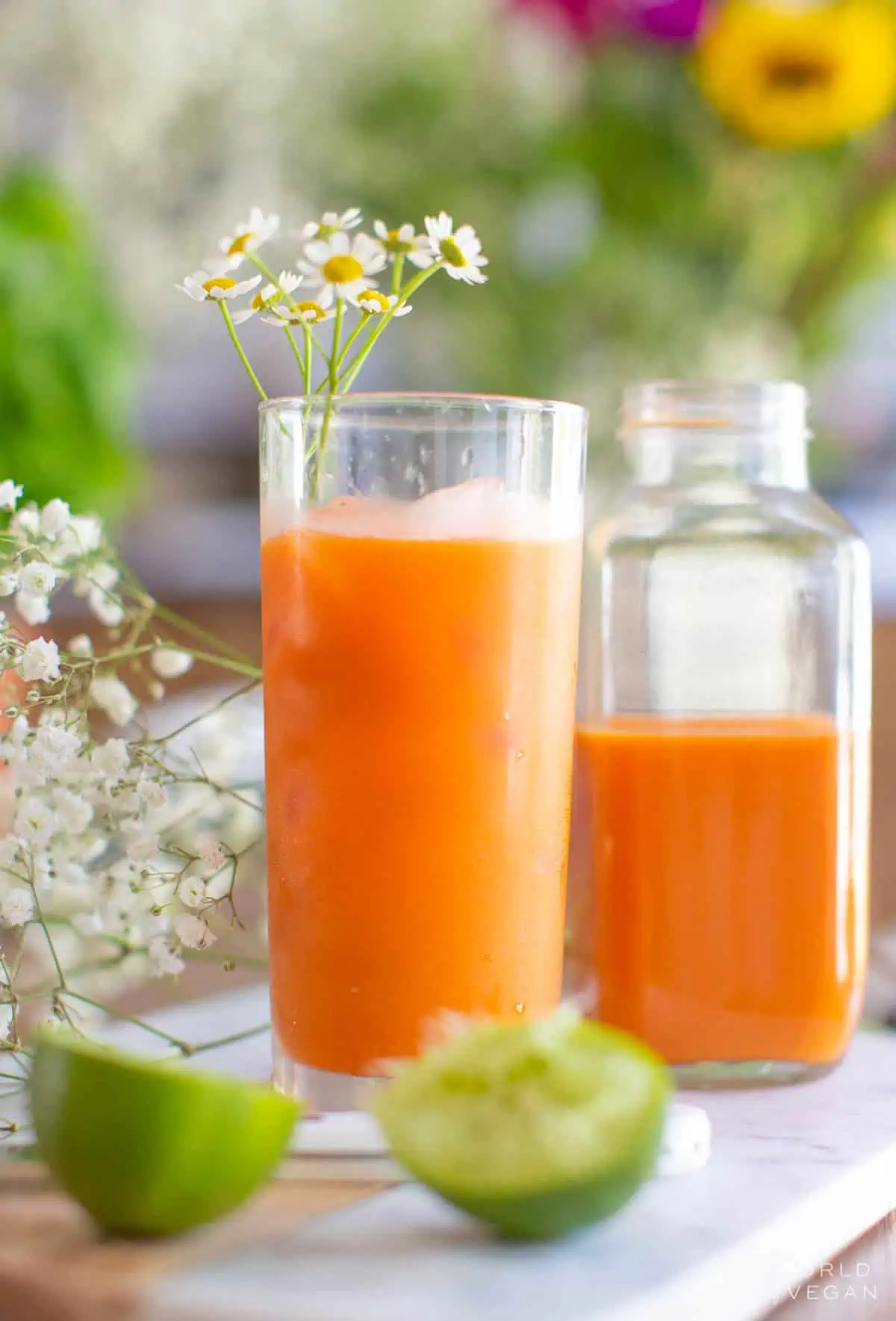 Tropical carrot juice in a glass next to a jar of carrot juice with lime wedges and flowers beside it.