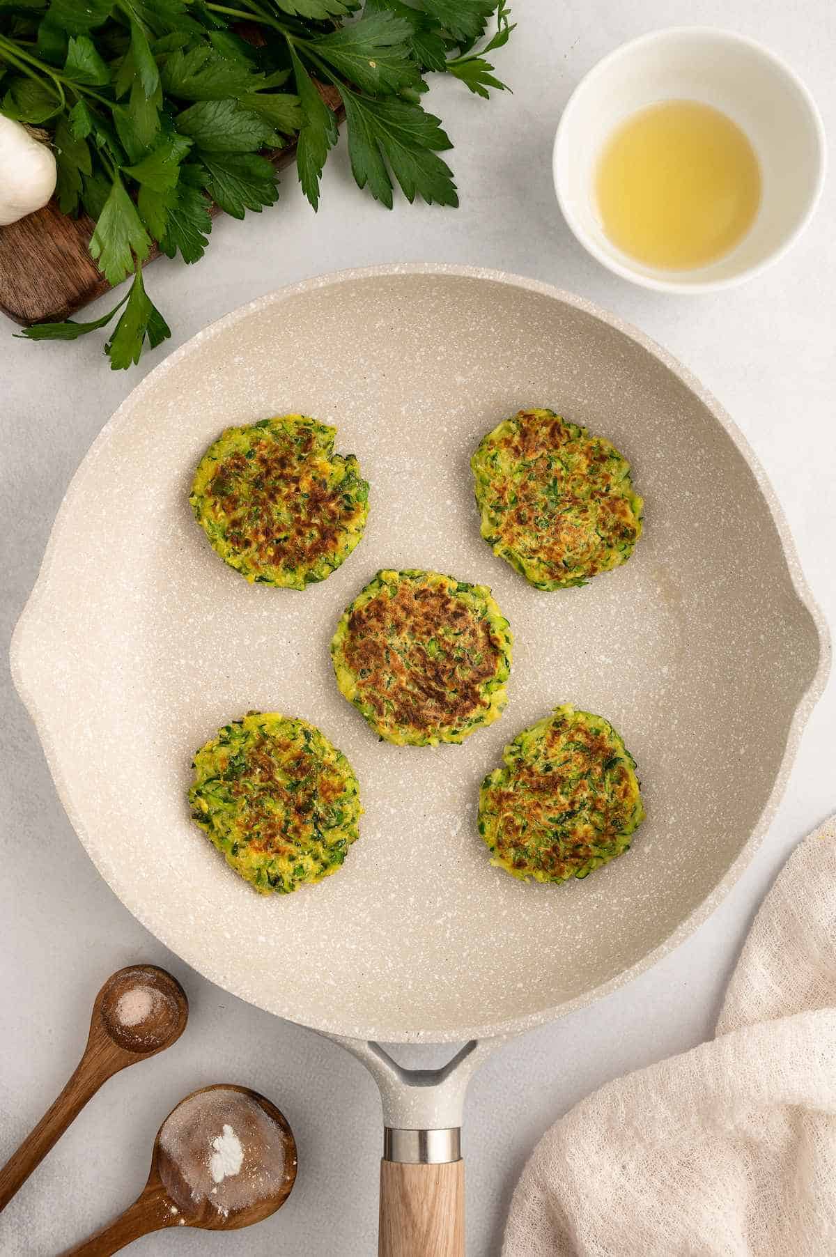 Five zucchini fritters in a pan that are cooked and golden.