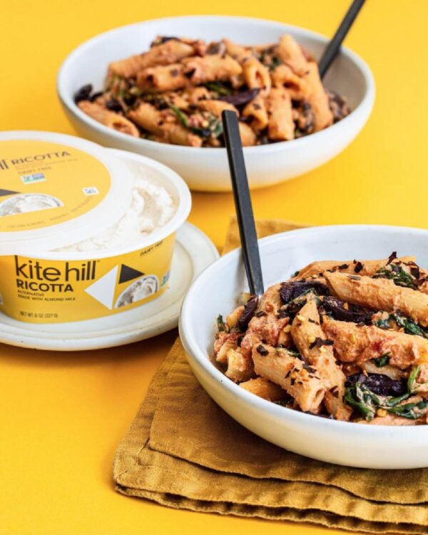 Two white bowls filled with pasta with spinach and vegan ricotta, alongside a small yellow tub of Kite Hill ricotta cheese against a yellow background.
