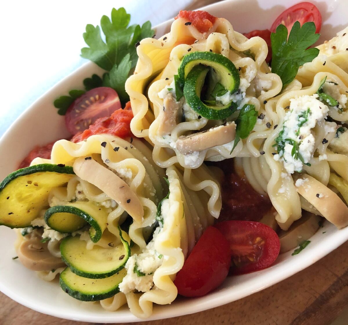 A large white bowl of rolled lasagna noodles, zucchini, tomatoes, and parsley along with Reine vegan cottage cheese against a white background. 