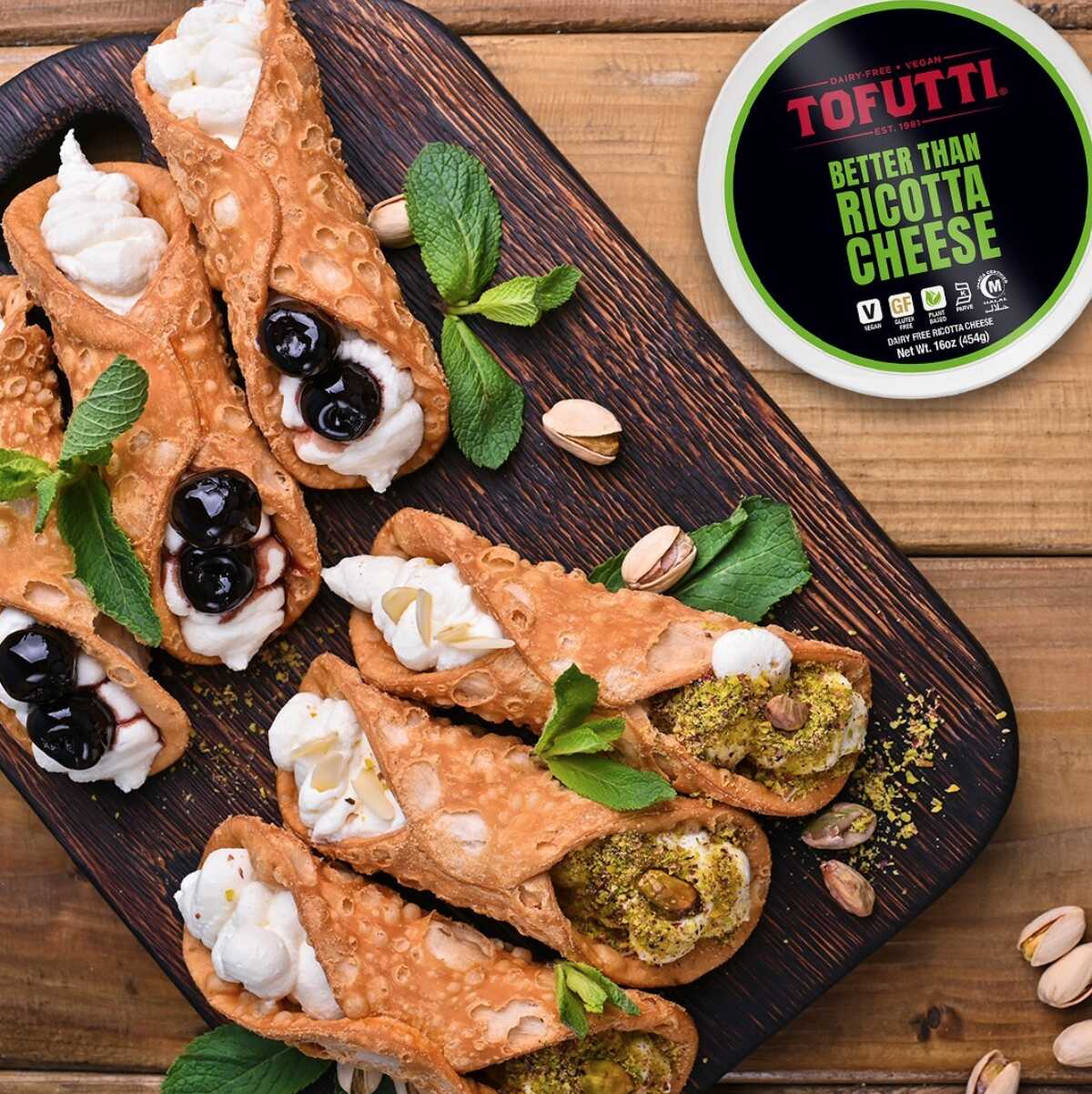 A dark wood cutting board topped with six cannolis that are filled with Tofutti vegan ricotta cheese. A black labeled plastic container of Tofutti ricotta is next to the cutting board. 