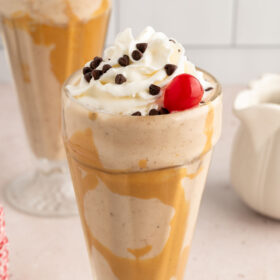 Vegan milkshake in a tall glass topped with vegan whipped cream, chocolate chips and a cherry.