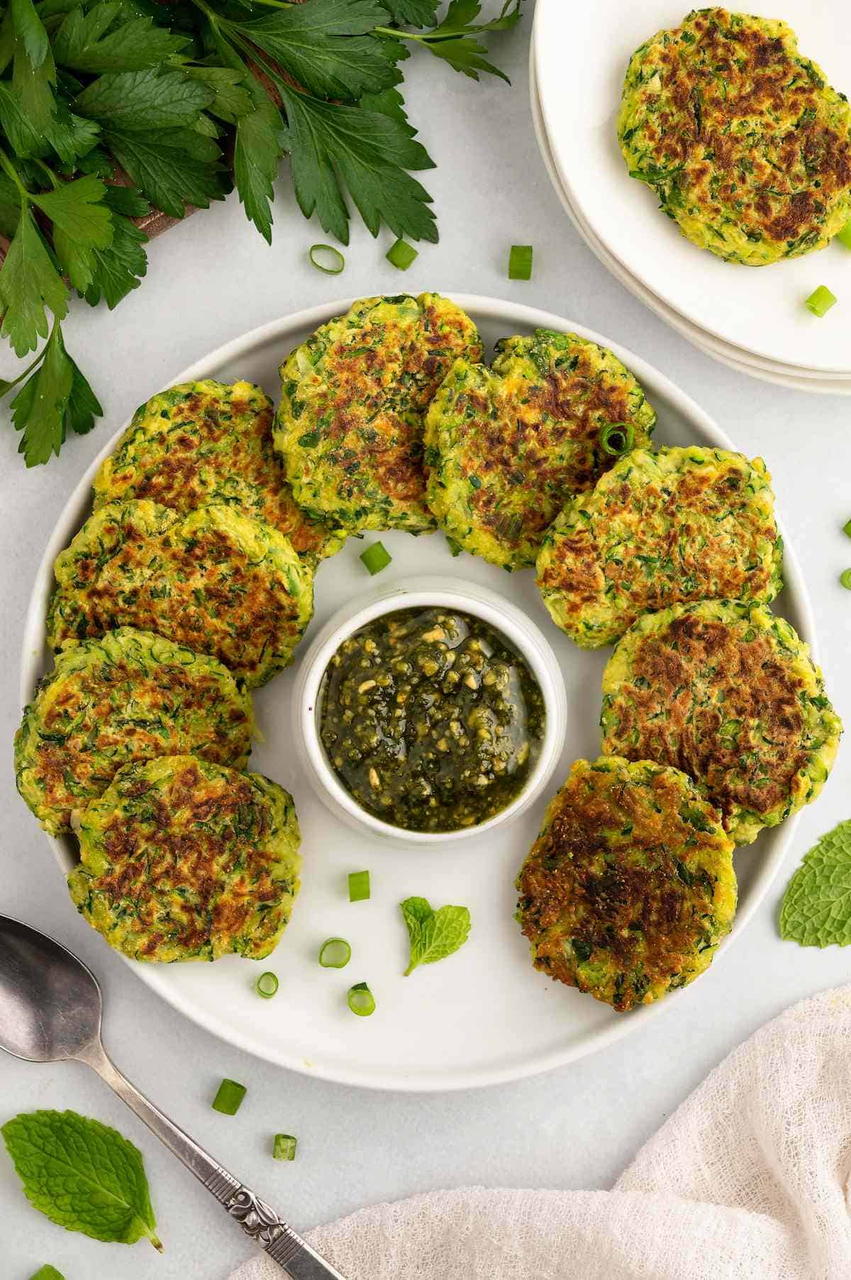 Vegan zucchini fritters placed in a circle around a small bowl of dip on a plate.