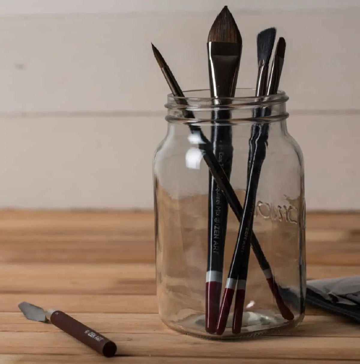 A glass jar filled with black paintbrushes on a wooden countertop.