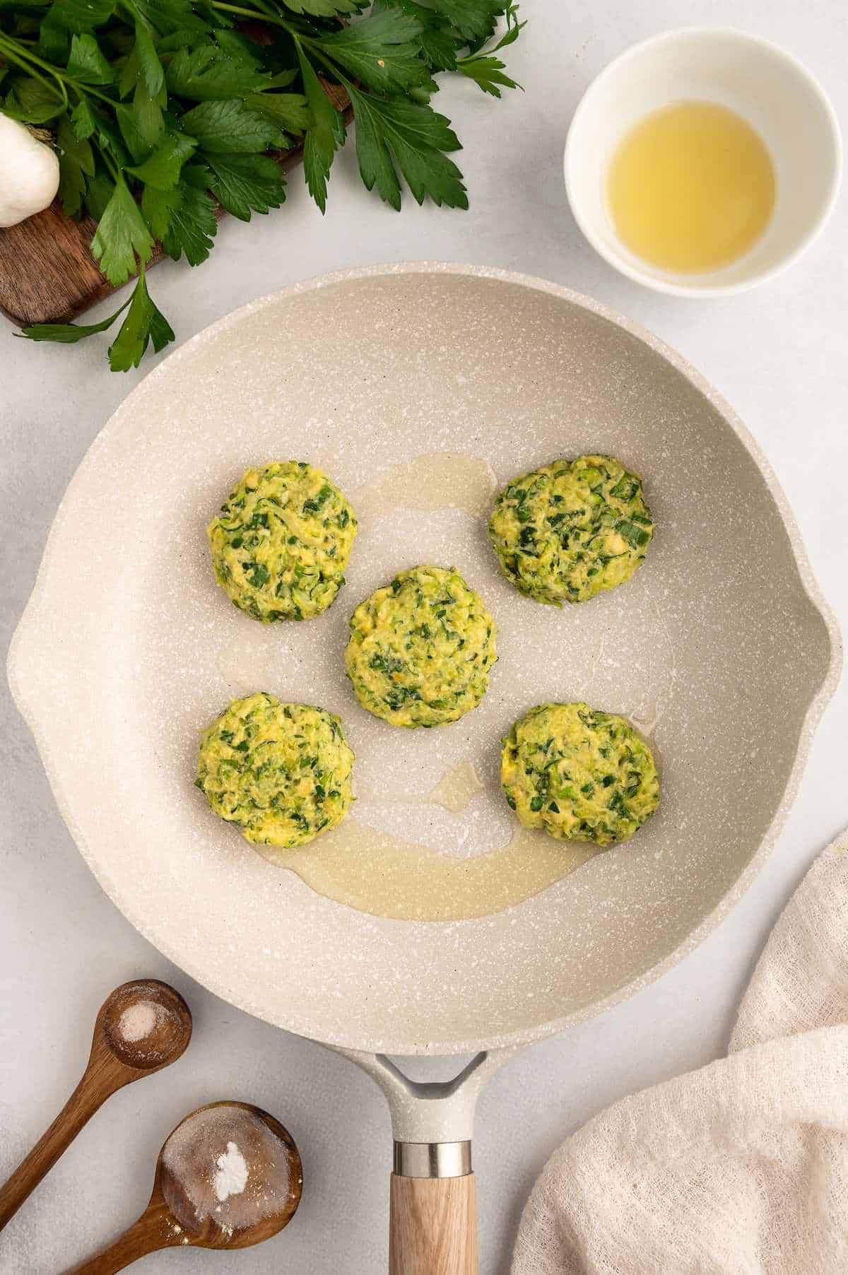 Five vegan zucchini fritters in a pan with some oil.