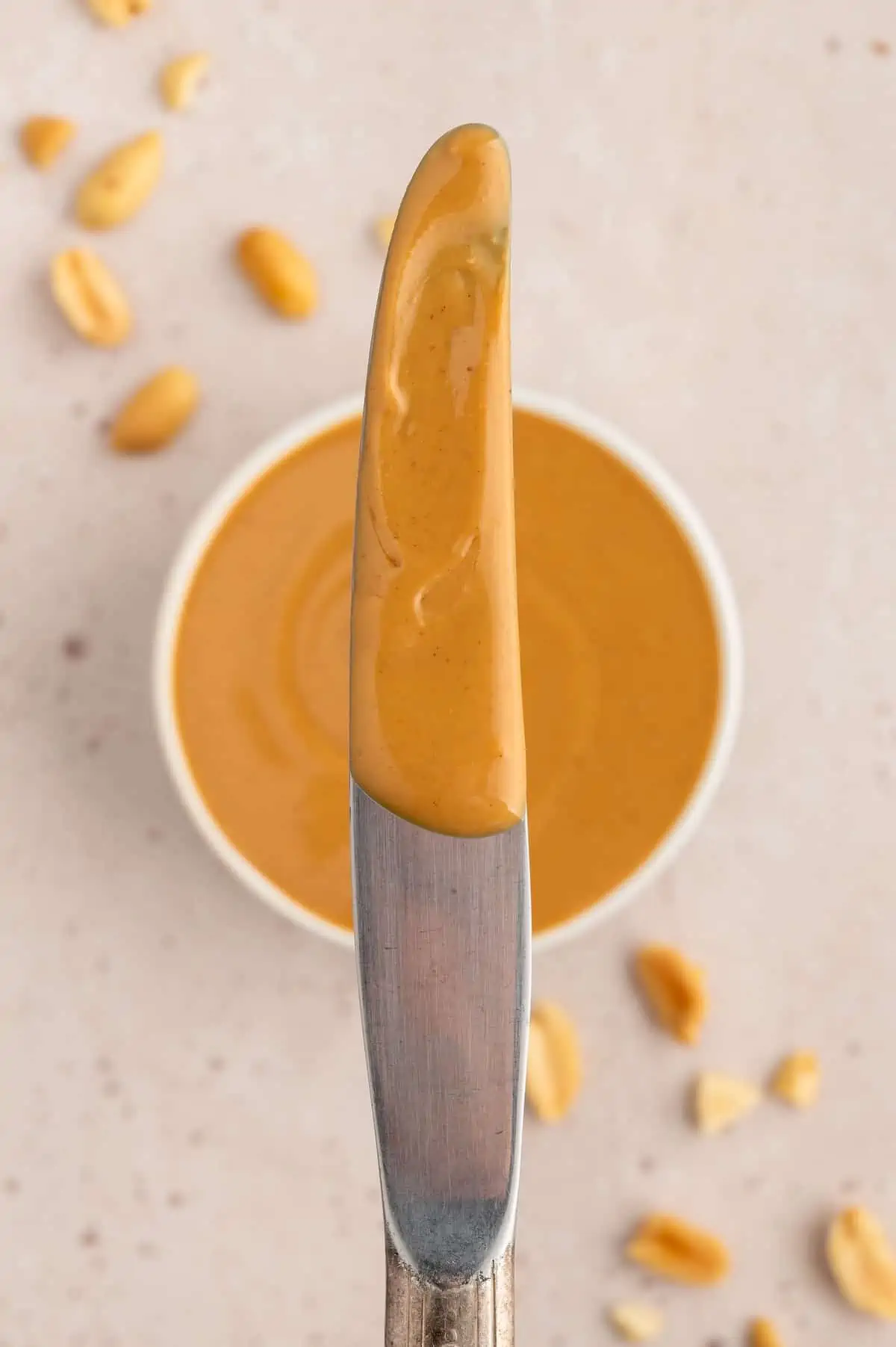 Smooth creamy peanut butter on a bread knife over a bowl of vegan peanut butter.