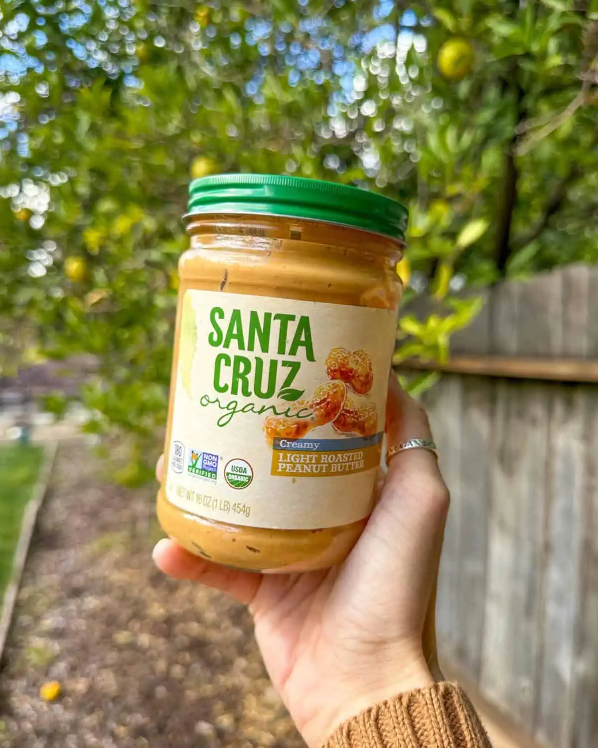 Woman's hand holding out a jar of Santa Cruz Organic creamy vegan peanut butter. 