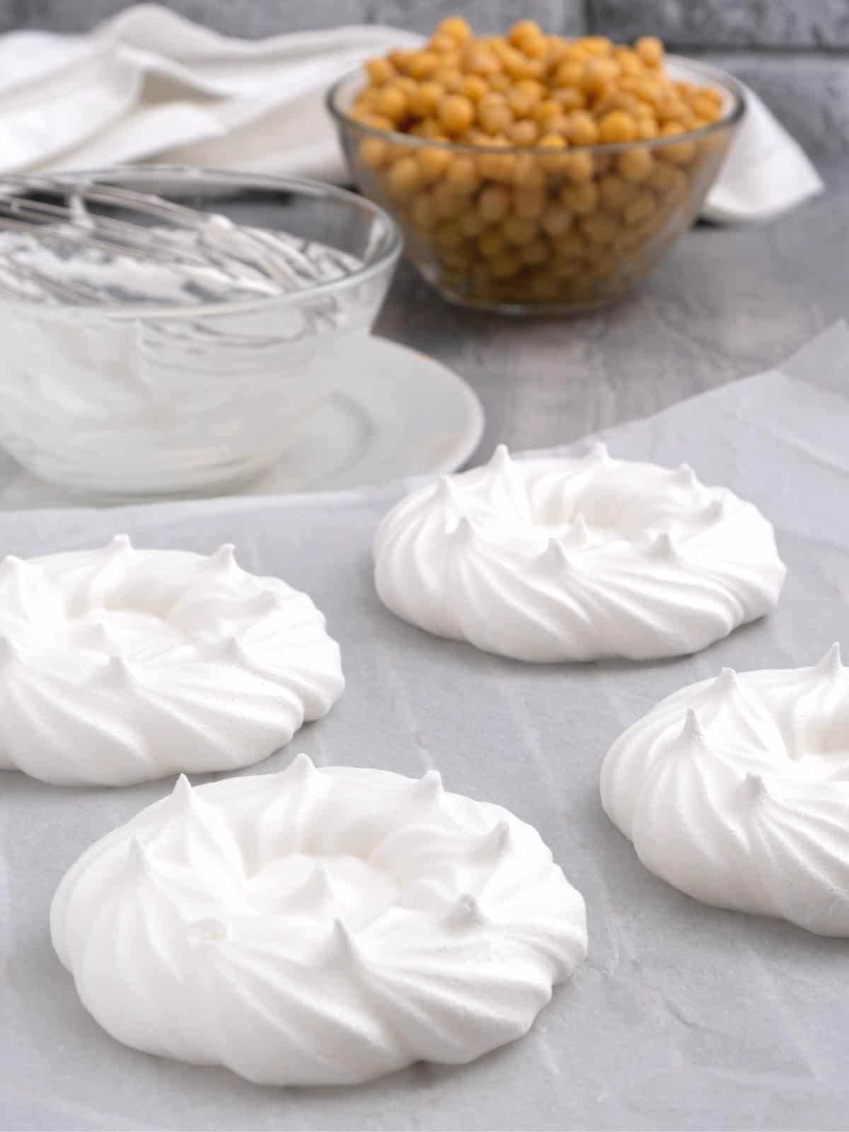 Vegan meringue cookies on parchment paper with a bowl of chickpeas in the background.