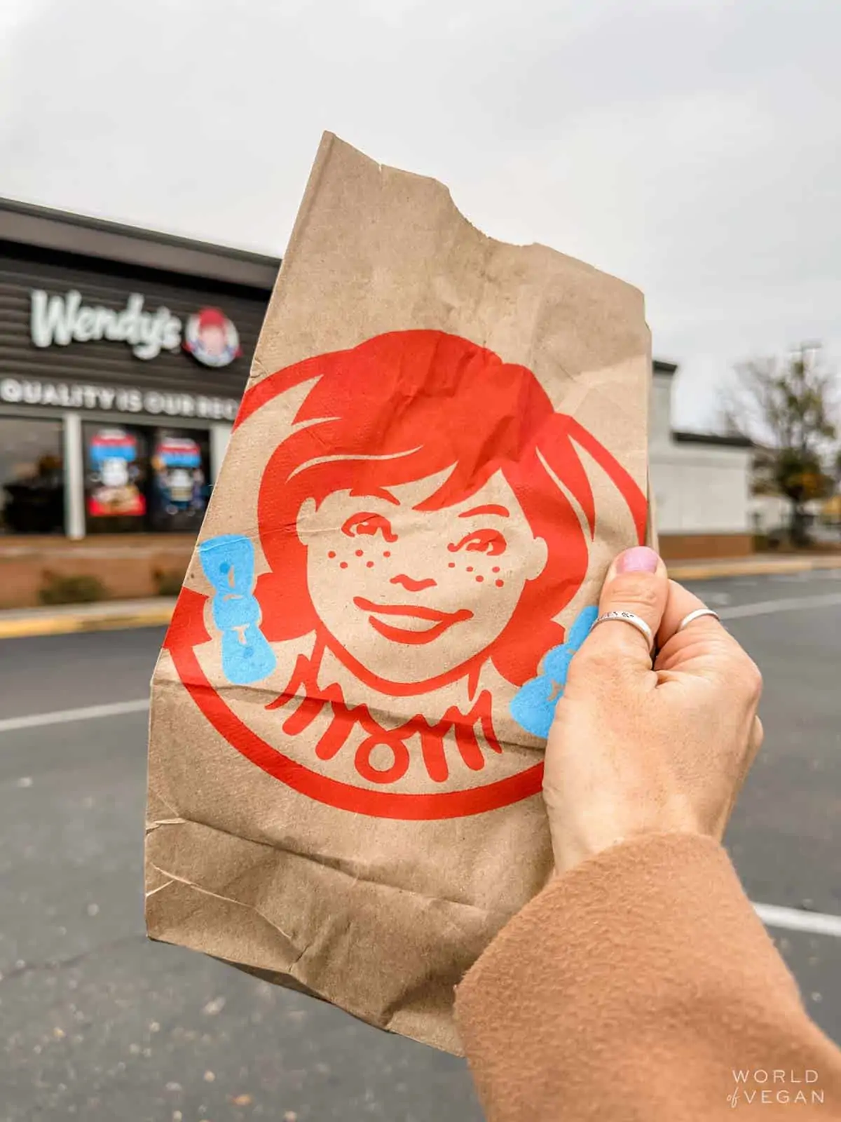 Holding up a Wendys paper bag in front of the Wendys restaurant.