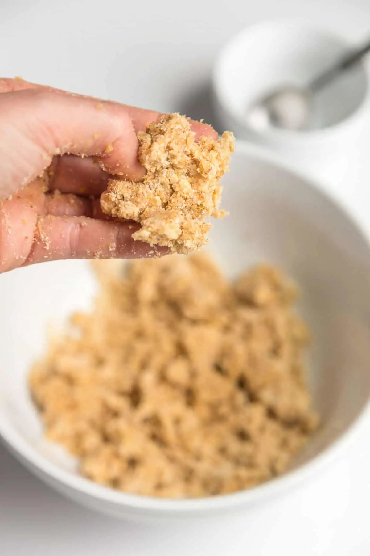 A bowl of almond flour cracker dough in a white bowl. One hand is holding some of the dough above the bowl.