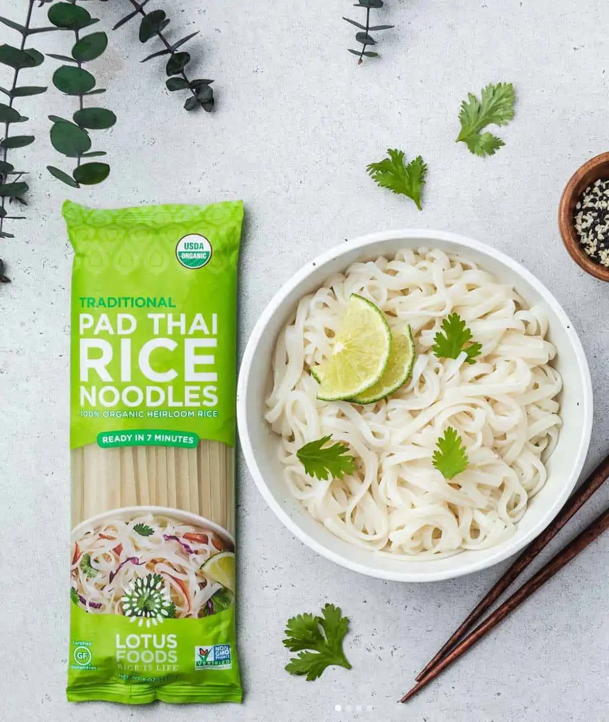 Lotus Foods Pad Thai Rice Noodles package next to a cooked bowl of rice noodles topped with cilantro and lime wedges.