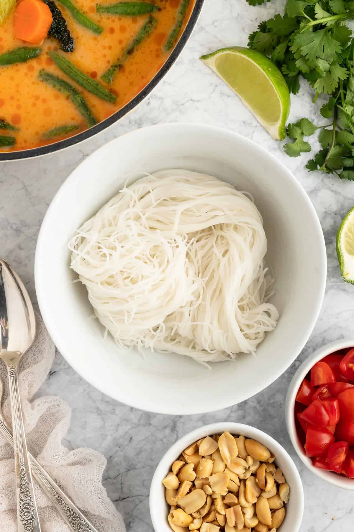 Rice vermicelli noodles in a white bowl, surrounded by herbs, peanuts and Thai curry broth in a pot to the left.