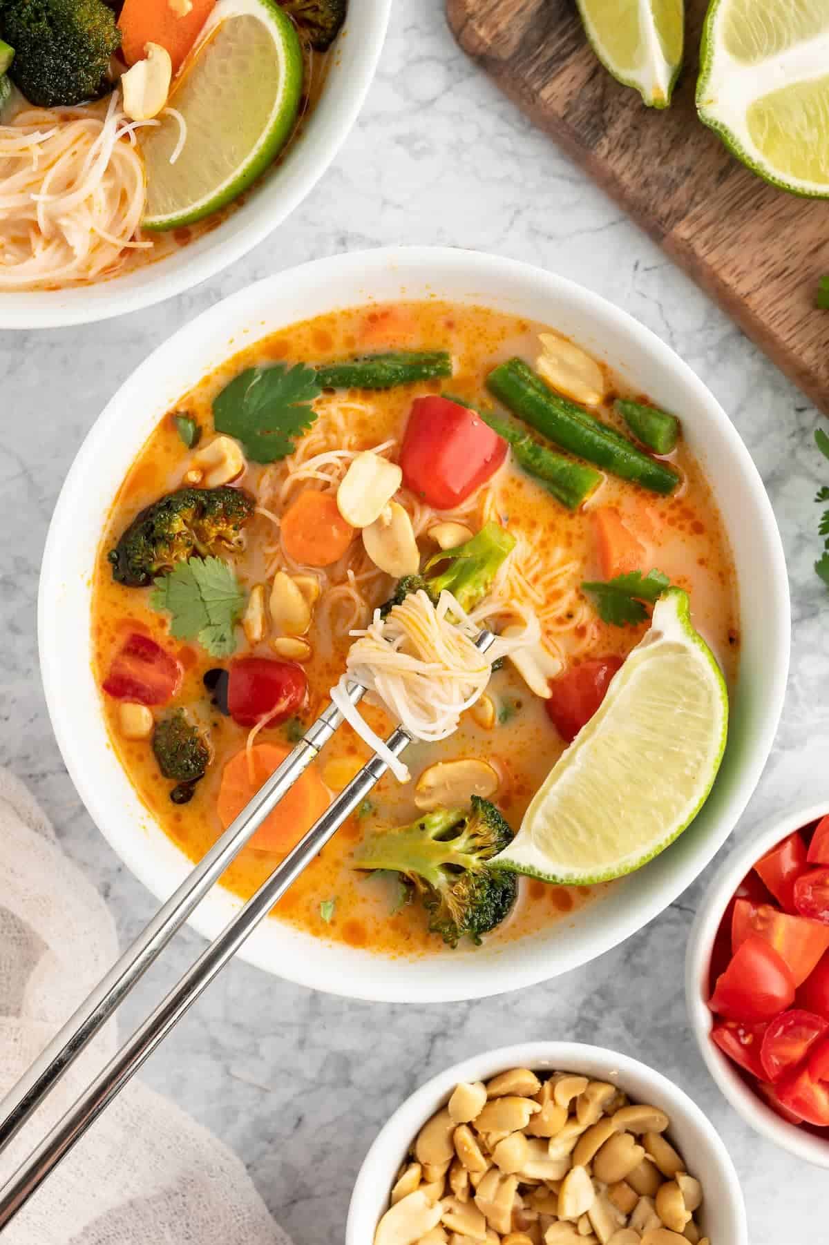 Thai curry in a white bowl with rice vermicelli wrapped around chopsticks. 