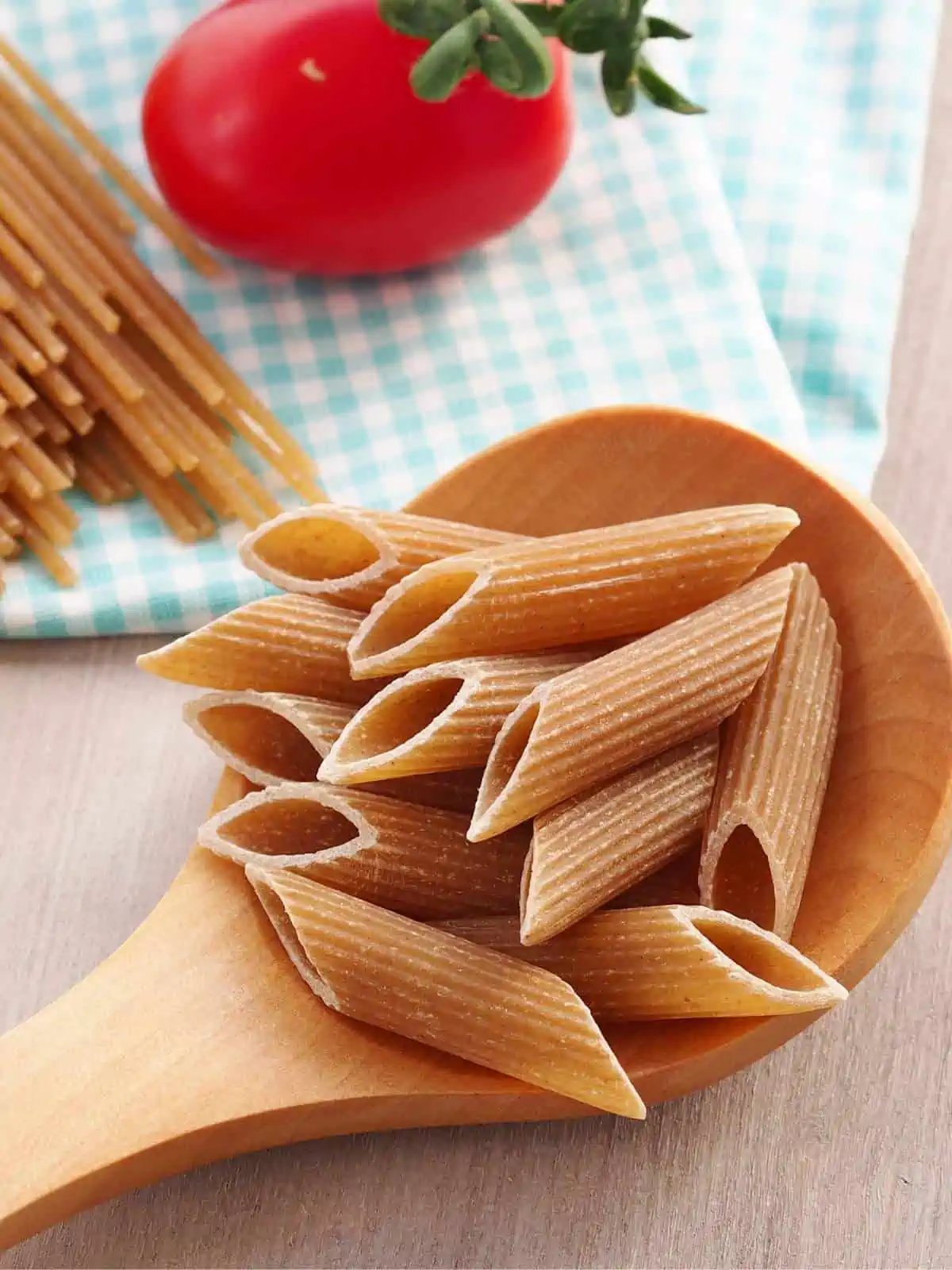Whole wheat penne pasta on a wooden spoon with whole spaghetti and a tomato behind it.