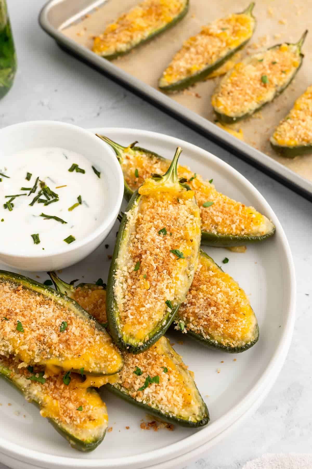 A plate of jalapeno poppers with dip on a white plate, with more poppers behind it on a baking sheet.