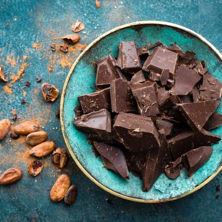 Broken pieces of a vegan dark chocolate block on a blue green plate with cacao beans and cocoa powder next to it.