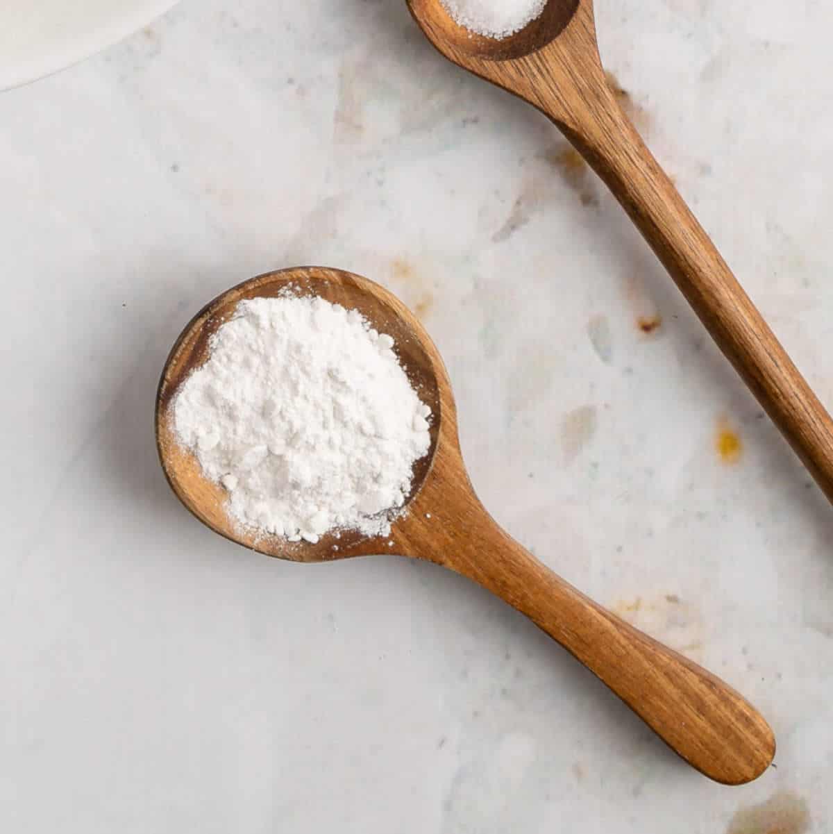 Two wooden tablespoons filled with baking ingredients on a light background.