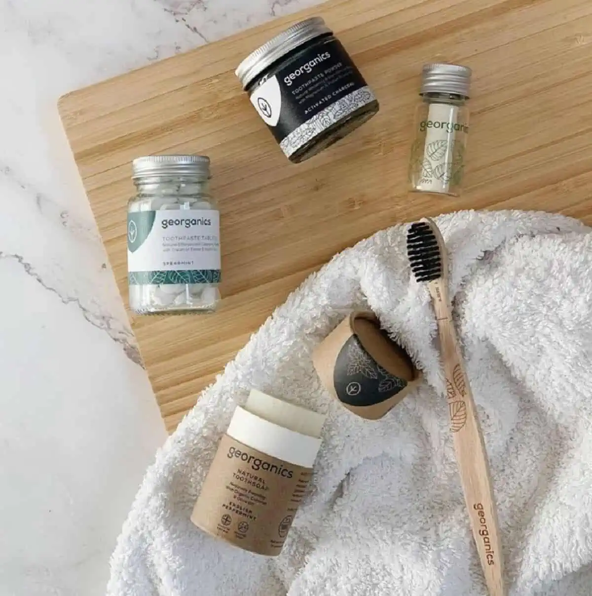 Three bottles of Georganics toothpaste tabs, mouthwash tabs and floss on a wooden cutting board next to toothpowder and a wooden tooth brush on a towel. 