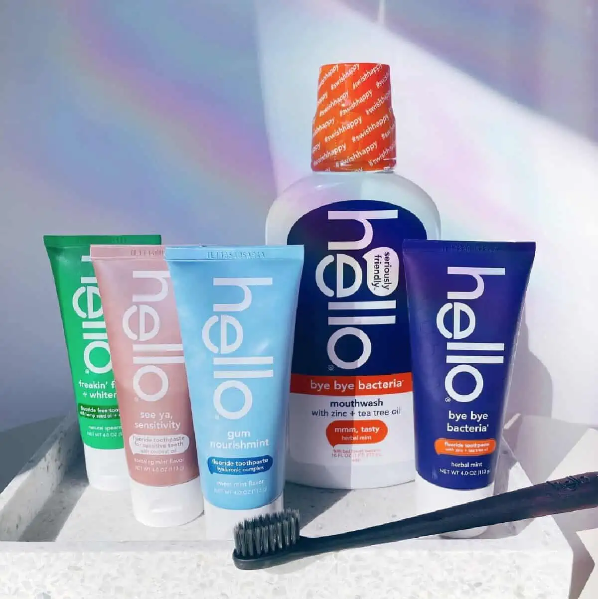 Four Hello toothpaste tubes, a bottle of Hello mouthwash and a black toothbrush against a rainbow-splashed white background. 