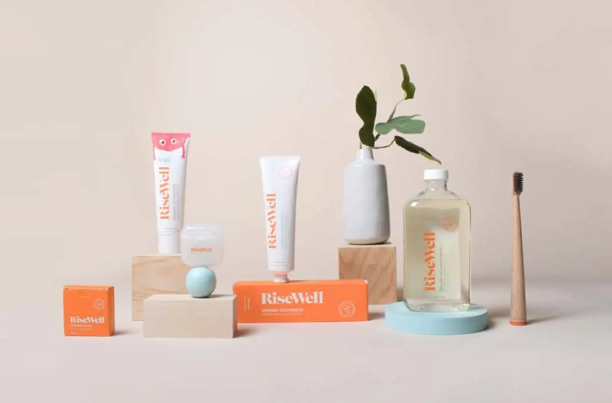 Two tubes of Risewell toothpaste, a container of floss and a bottle of mouthwash on wooden blocks set against a beige background with a small plant in a vase.