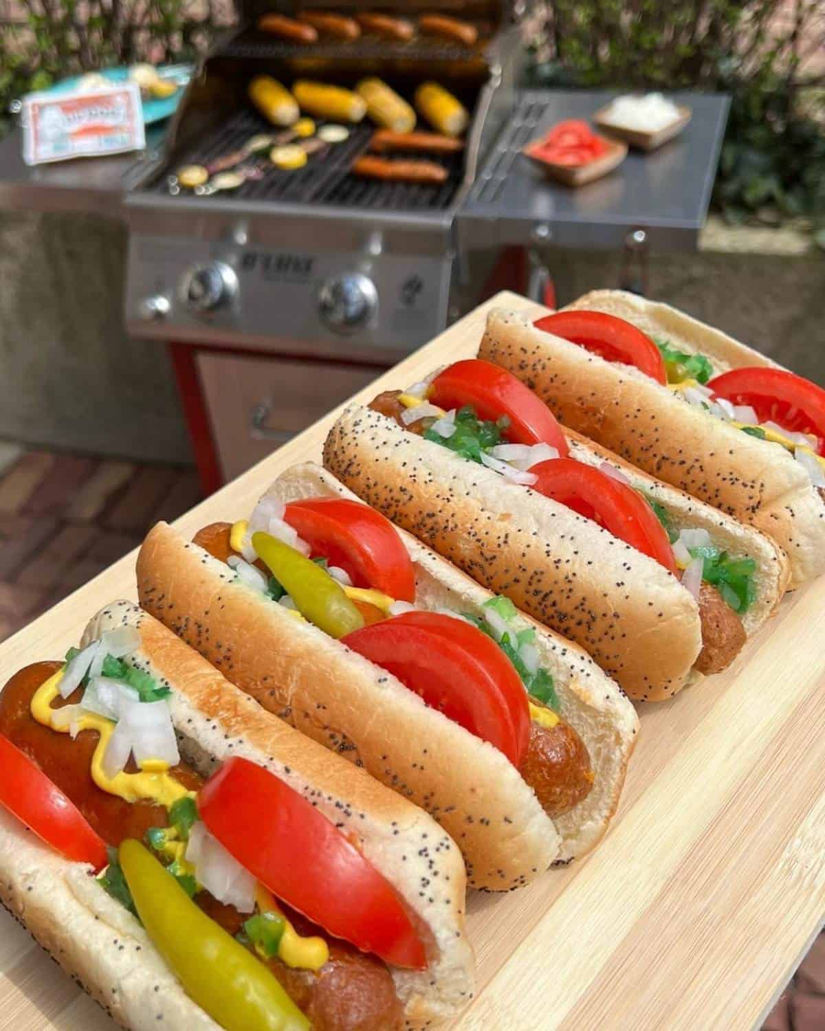 Four Upton's hot dogs in buns with toppings and a grill in the background.