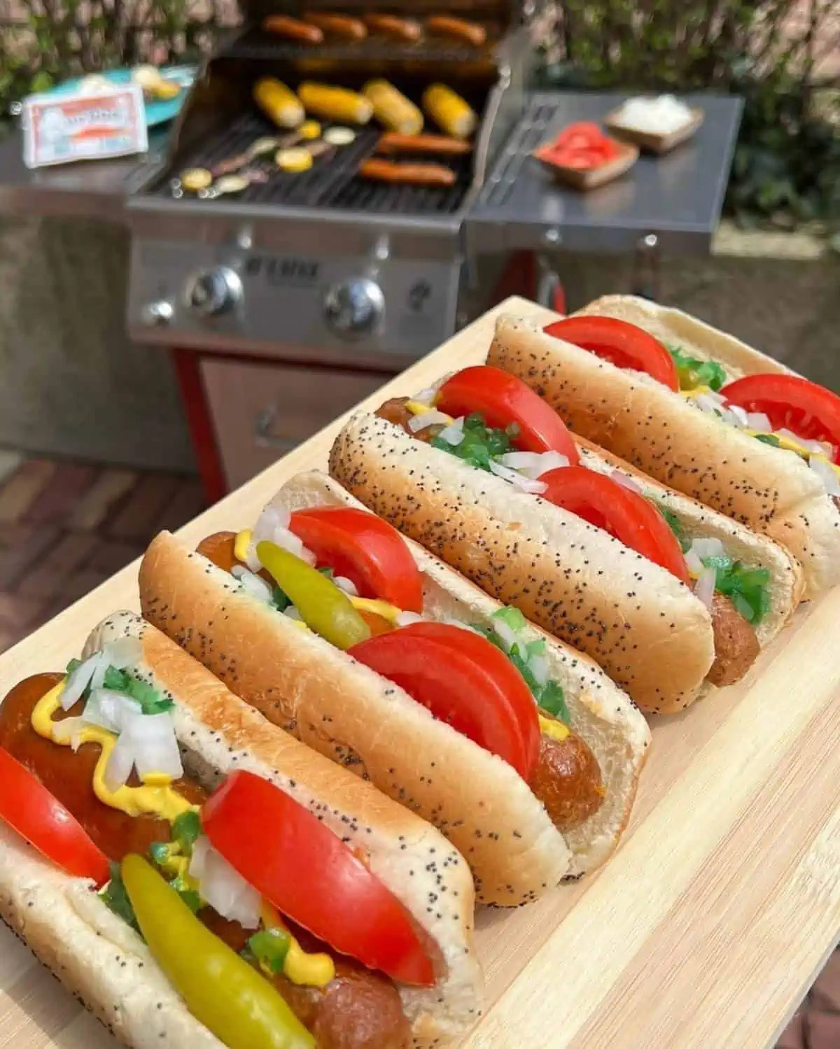 Four Upton's hot dogs in buns with toppings and a grill in the background.