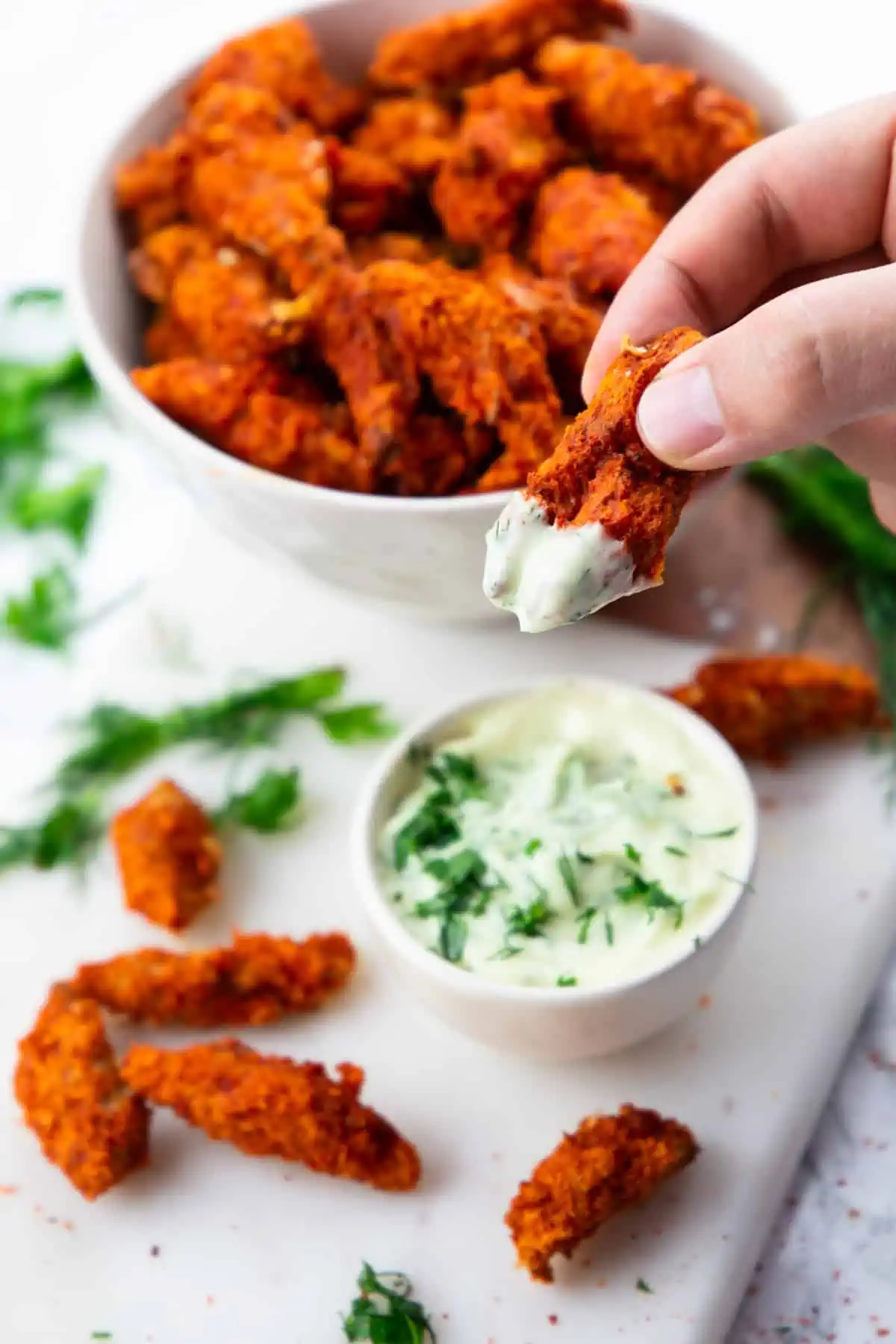 A bowl of vegan chicken behind a hand holding one piece of vegan chicken that has been dipped in vegan ranch sauce.