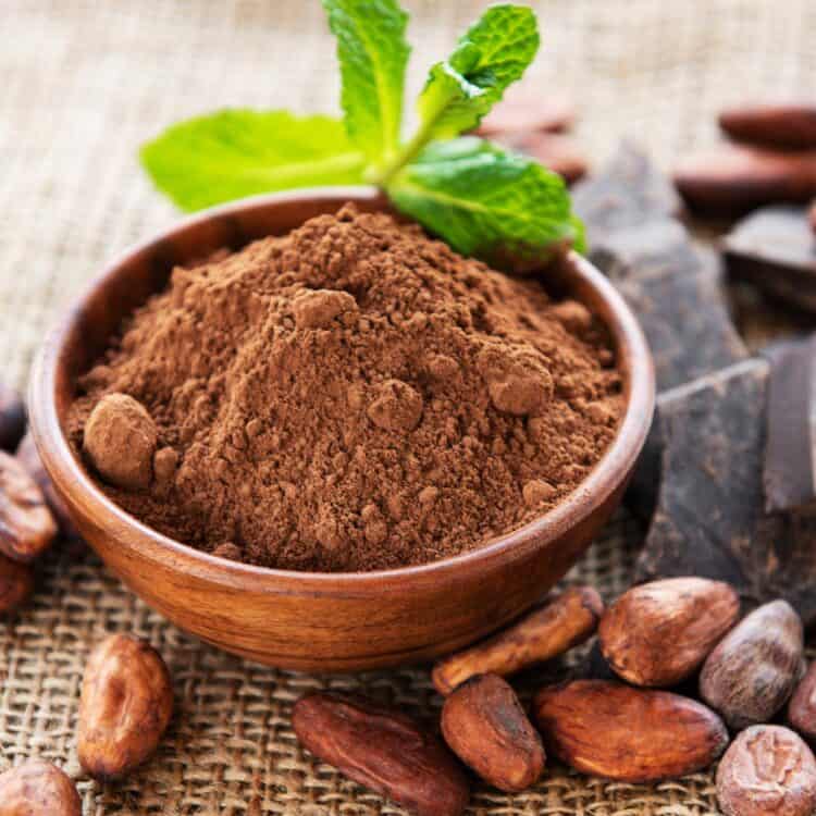 Wooden bowl of cocoa powder surrounded by cacao beans and dark chocolate.