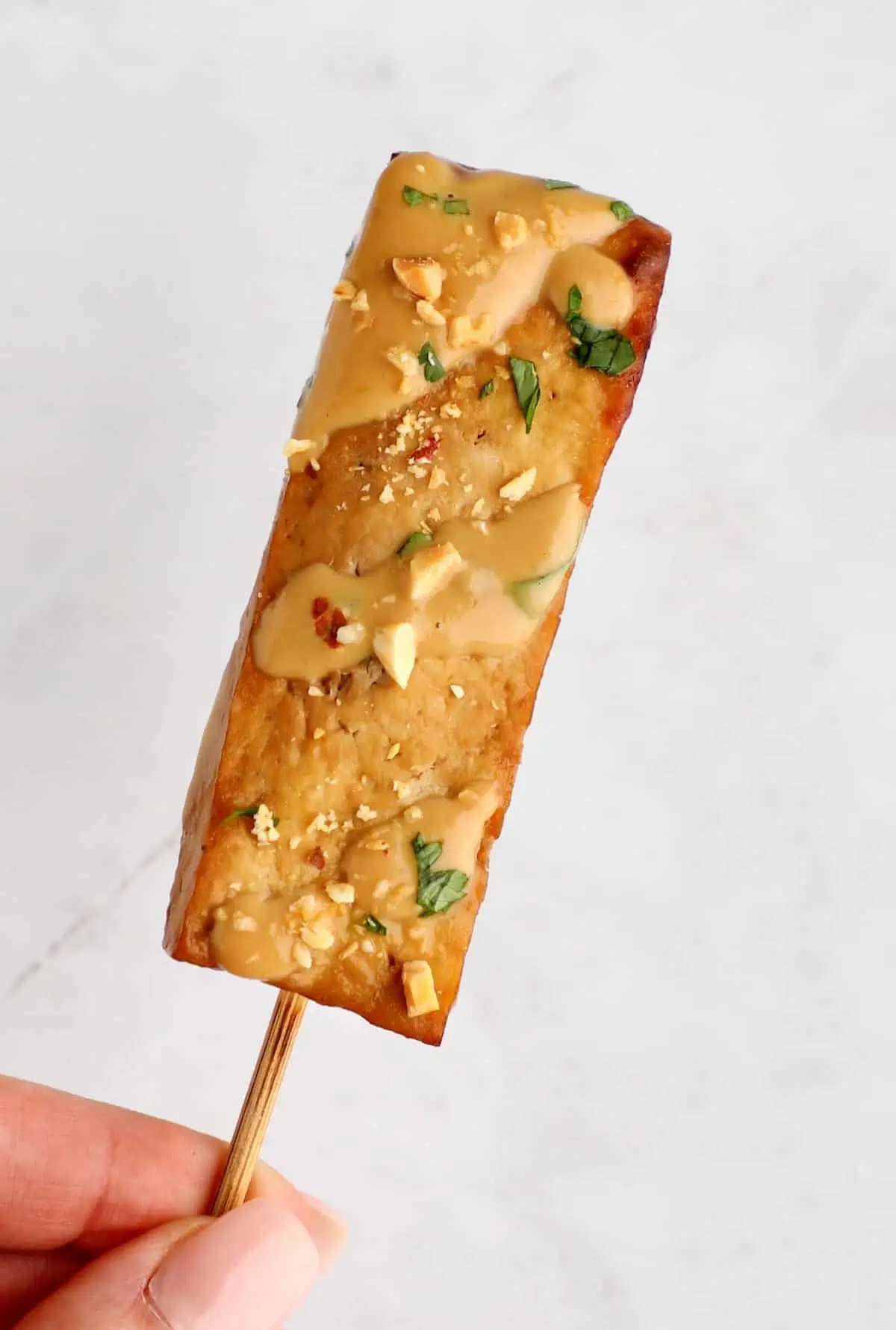 Hand holding one tofu satay skewer that's been drizzle with peanut dressing.