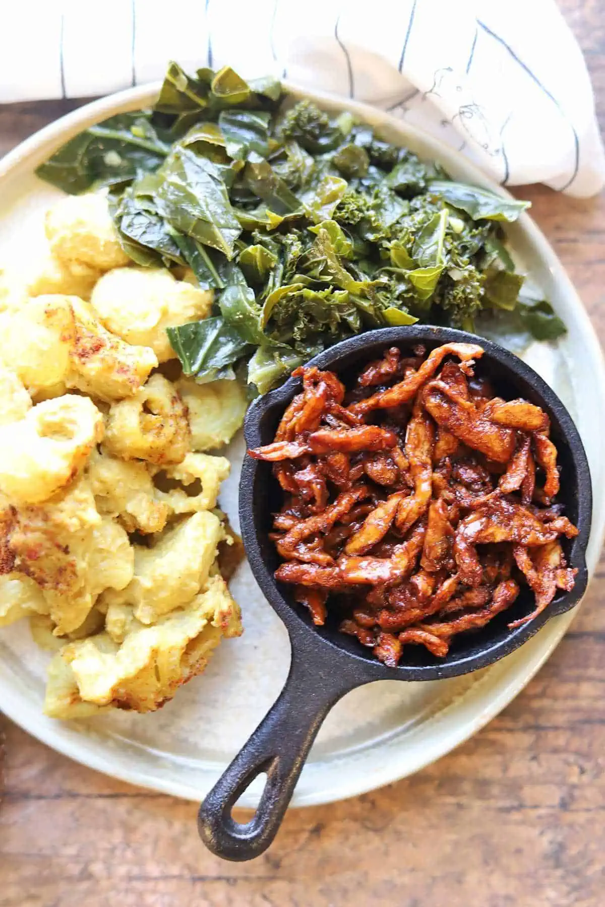 A small cast iron skillet filled with vegan pulled pork positioned on a plate next to cooked greens and cauliflower.