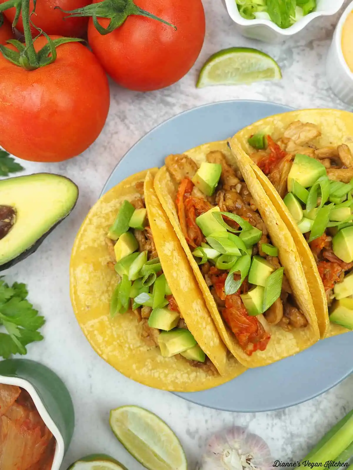 Three soy curl kimchi tacos on a plate surrounded by various fresh ingredients.