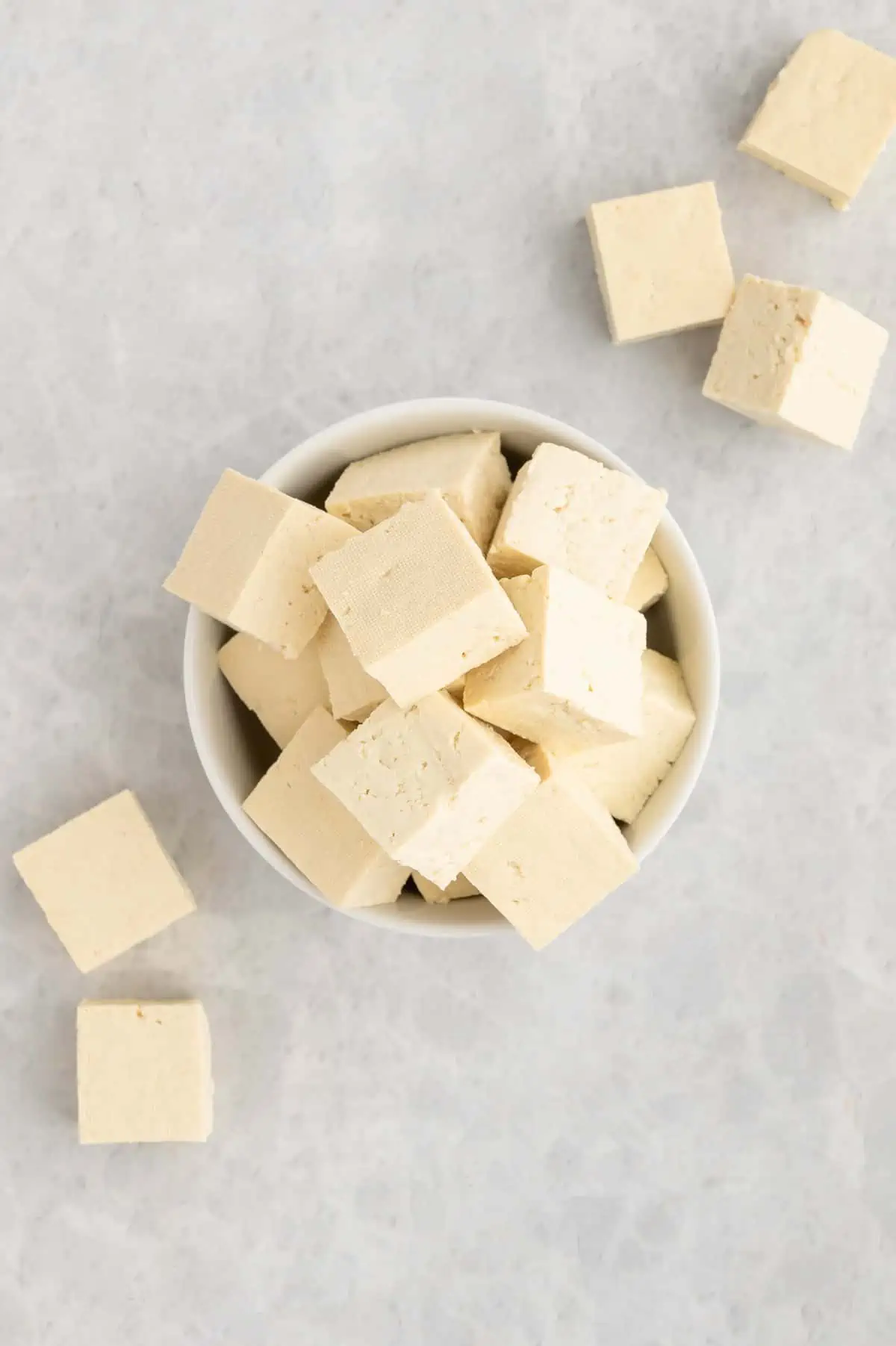 Small bowl full of raw tofu cubes with some cubes scattered around the bowl.