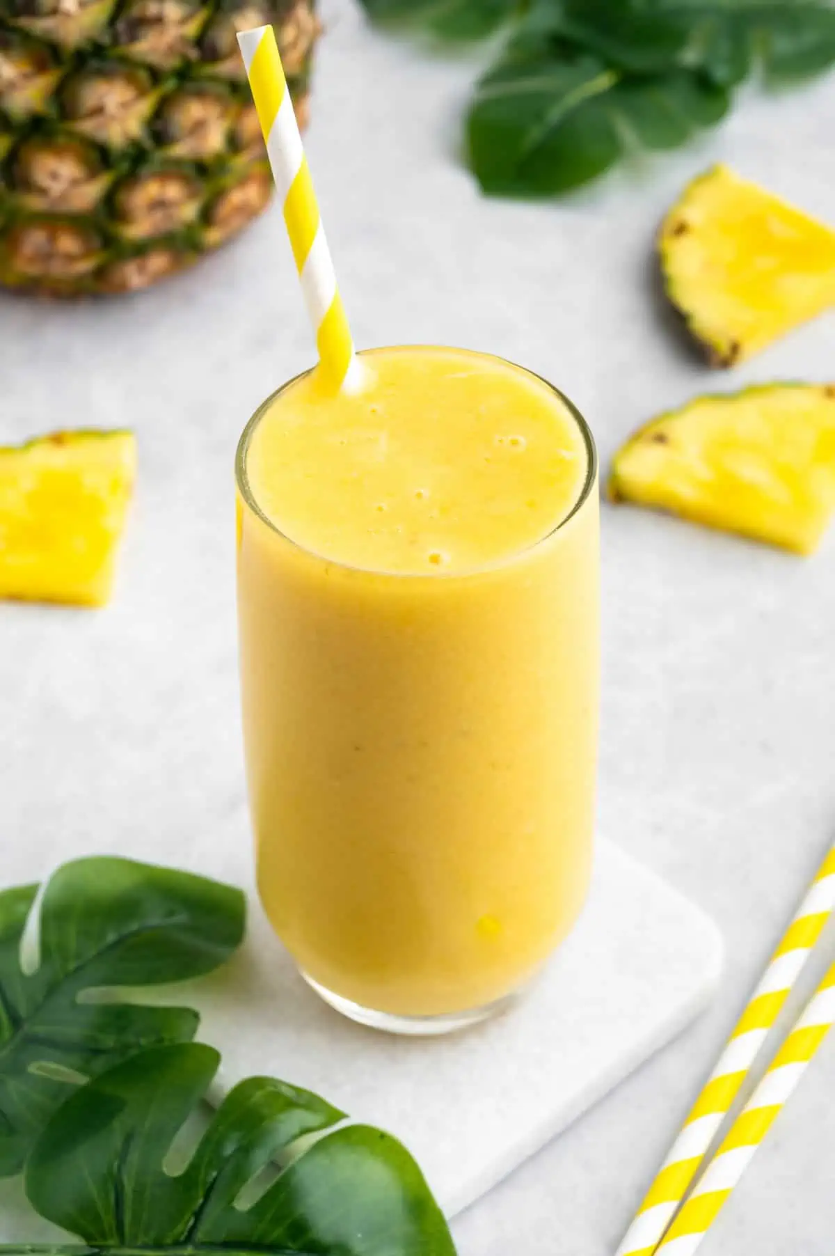Mango pineapple smoothie in a glass with a straw, surround by slices of pineapple.