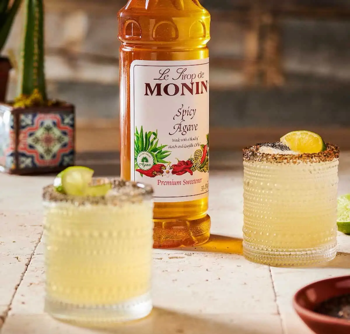 Bottle of Le Sirop de Monin Spicy Agave next to two small glasses of margaritas with limes.
