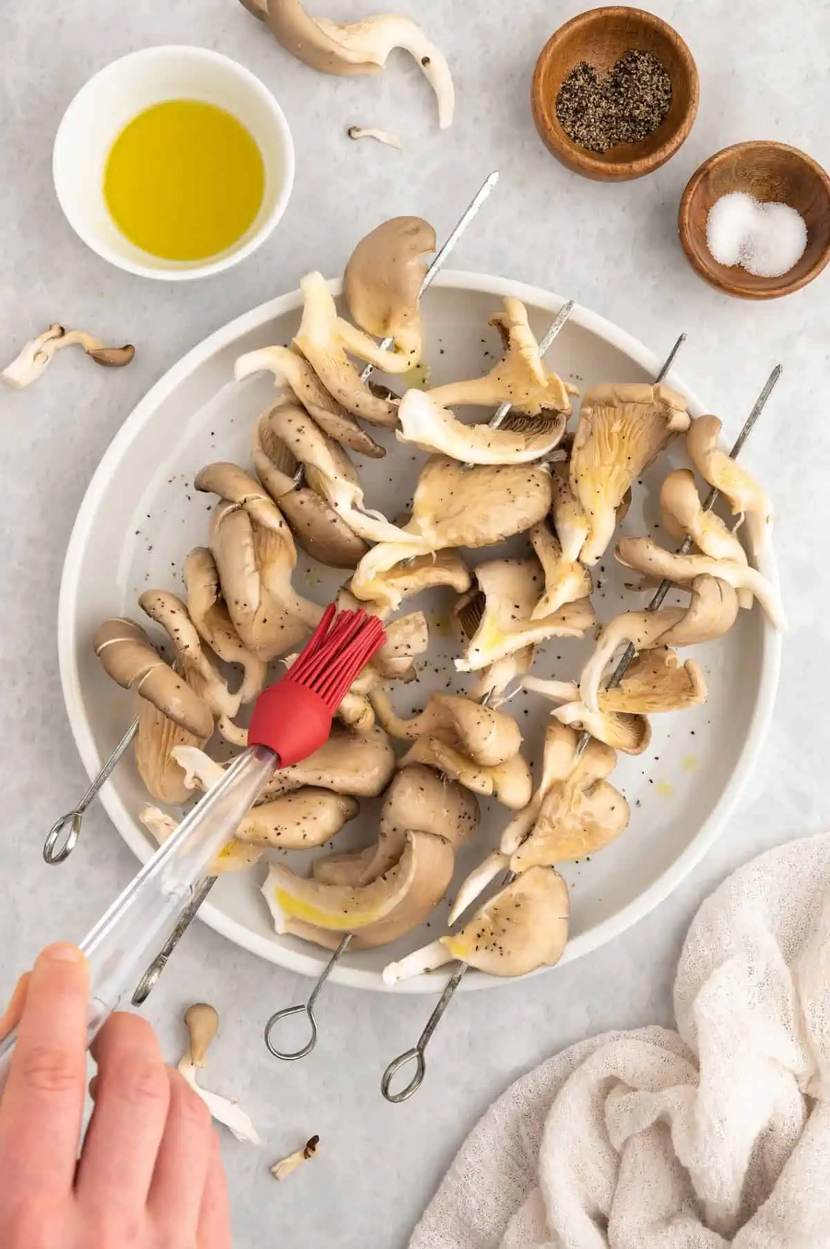 Oyster mushroom threaded onto metal skewers being brushed with olive oil.