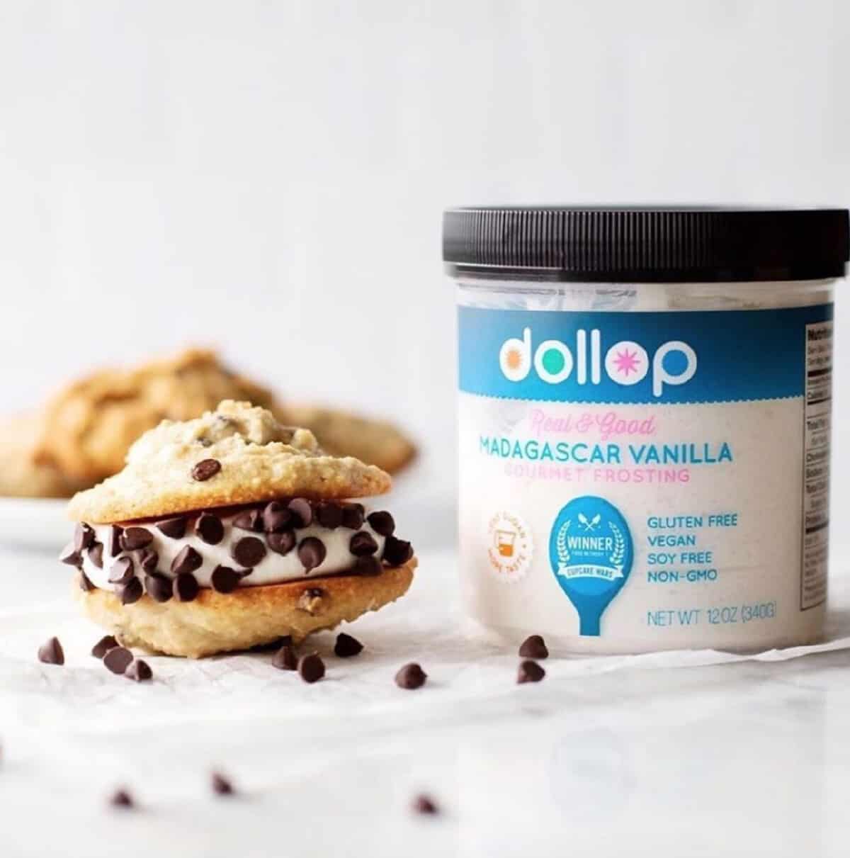 A can of Dollop vanilla vegan frosting on a white table next to a chocolate chip cookie sandwich. 