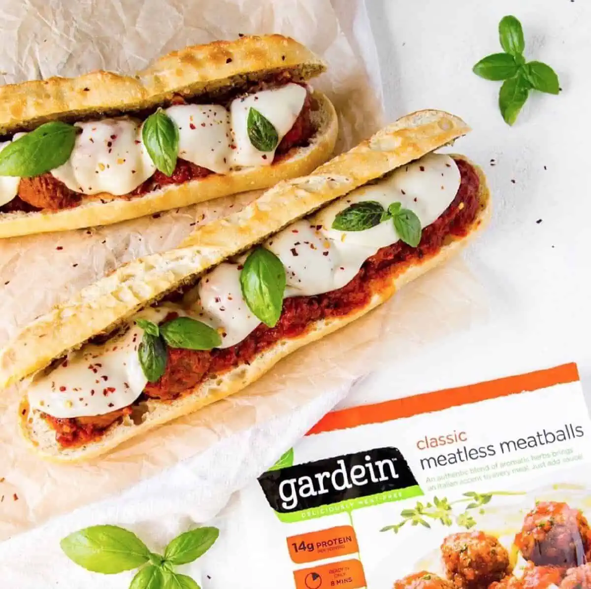 Two large vegan meatballs subs with Gardein meatballs, sauce, vegan cheese, and fresh basil leaves next to a bag of Gardein meatballs on a white background.