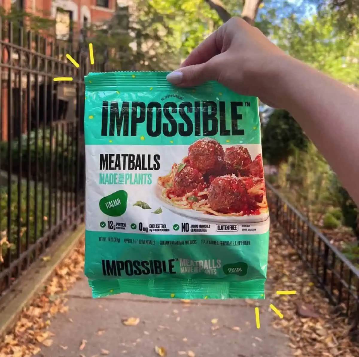 A hand holding a green and white bag of Impossible Meatballs on a city street with leaves on the ground.